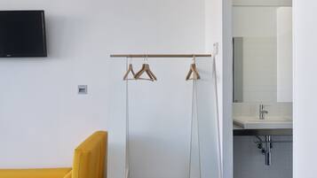A modern hotel room with a flat-screen TV mounted on the wall, a wooden coat rack with hangers, a yellow sofa, and a bathroom with a sink and mirror.