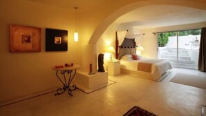 A bedroom with a bed, bedside tables, a small table, and a lamp. There is a window with curtains and a view of the outside. - Sitio Sagrado (Tepoztlán)