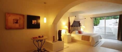 A bedroom with a bed, bedside tables, a small table, and a lamp. There is a window with curtains and a view of the outside.