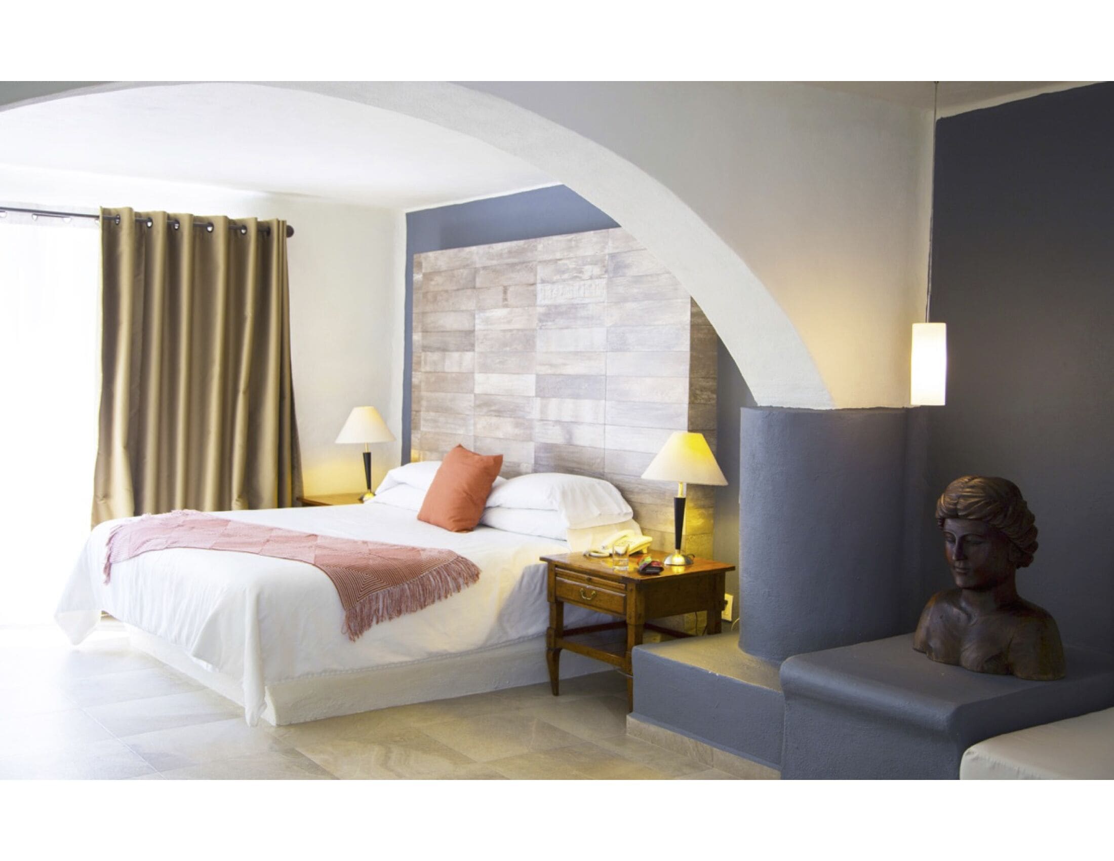 A hotel room with a bed, bedside tables, a lamp, a sculpture, and a stone wall.