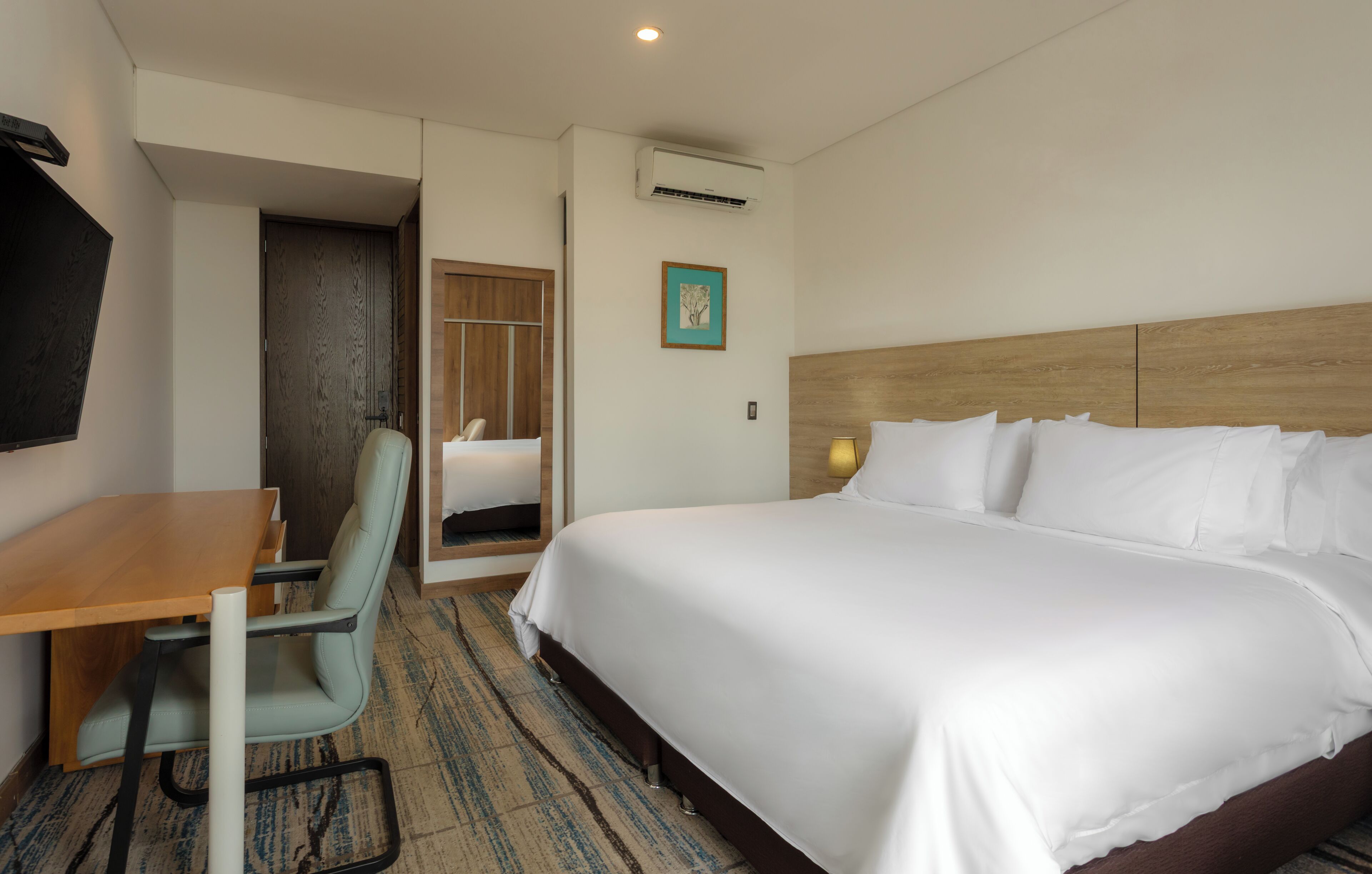 Une chambre d’hôtel équipée d’un lit, d’un bureau, d’une chaise, d’un climatiseur et d’un tableau encadré au mur.