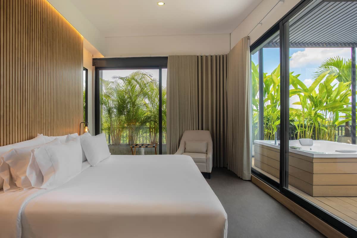 A modern hotel room with a large bed, a seating area with a chair, and a view of tropical plants through a large window.