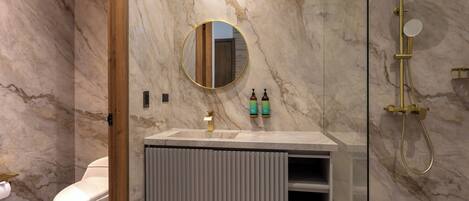 Un baño moderno con pared de mármol, un espejo redondo, inodoro, tocador con toallero y ducha con detalles dorados.