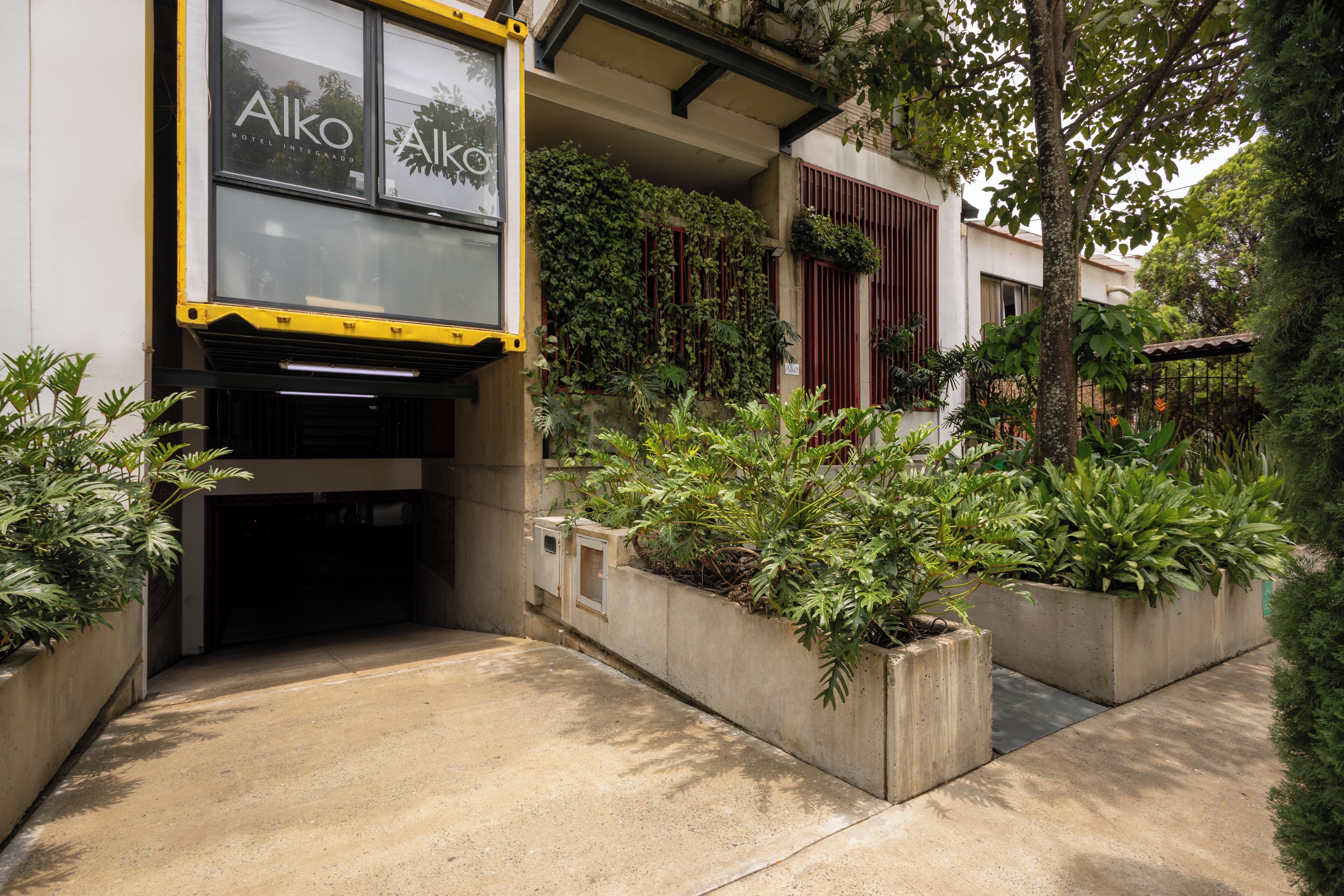 Fachada de hotel en amarillo y negro, macetas de concreto con plantas y una puerta visible.