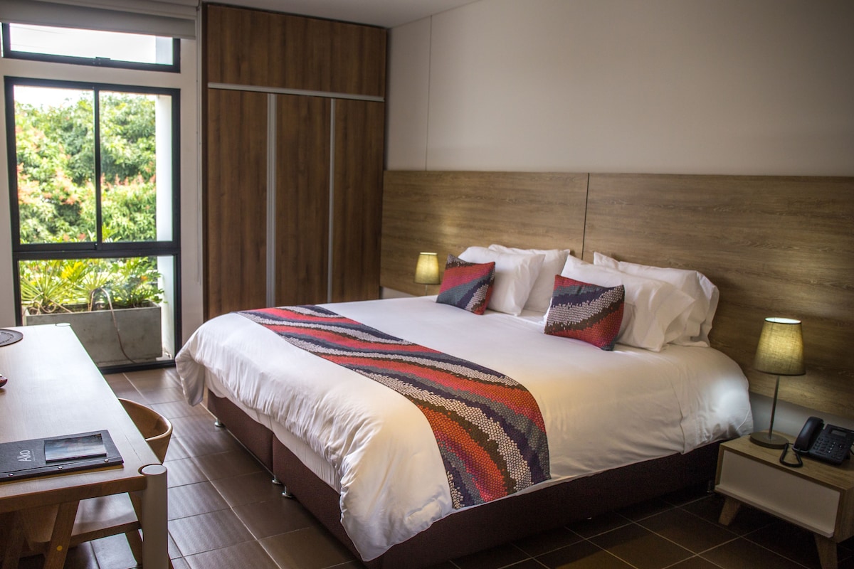 Ein Hotelzimmer mit einem großen Bett, Nachttischlampen, einem Schreibtisch und Blick auf Grünflächen.