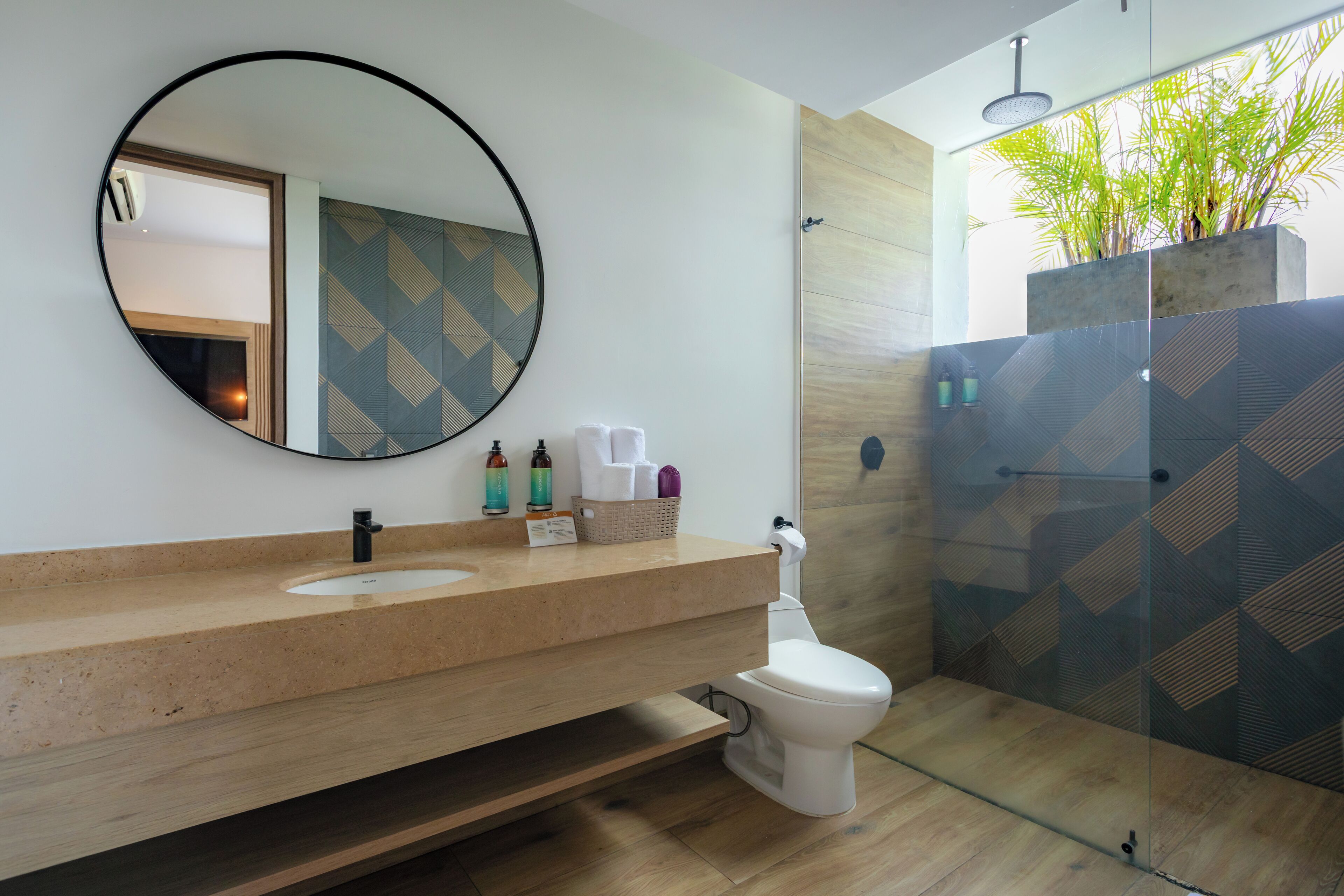 Un baño moderno con un espejo redondo, un lavabo y un inodoro. Cuenta con una zona de ducha con puerta de cristal y una planta en maceta.