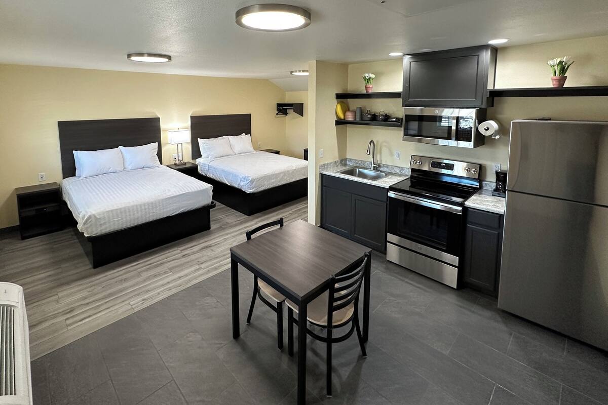 A compact hotel room with two beds, a small kitchenette, a dining table, and a window with blinds.