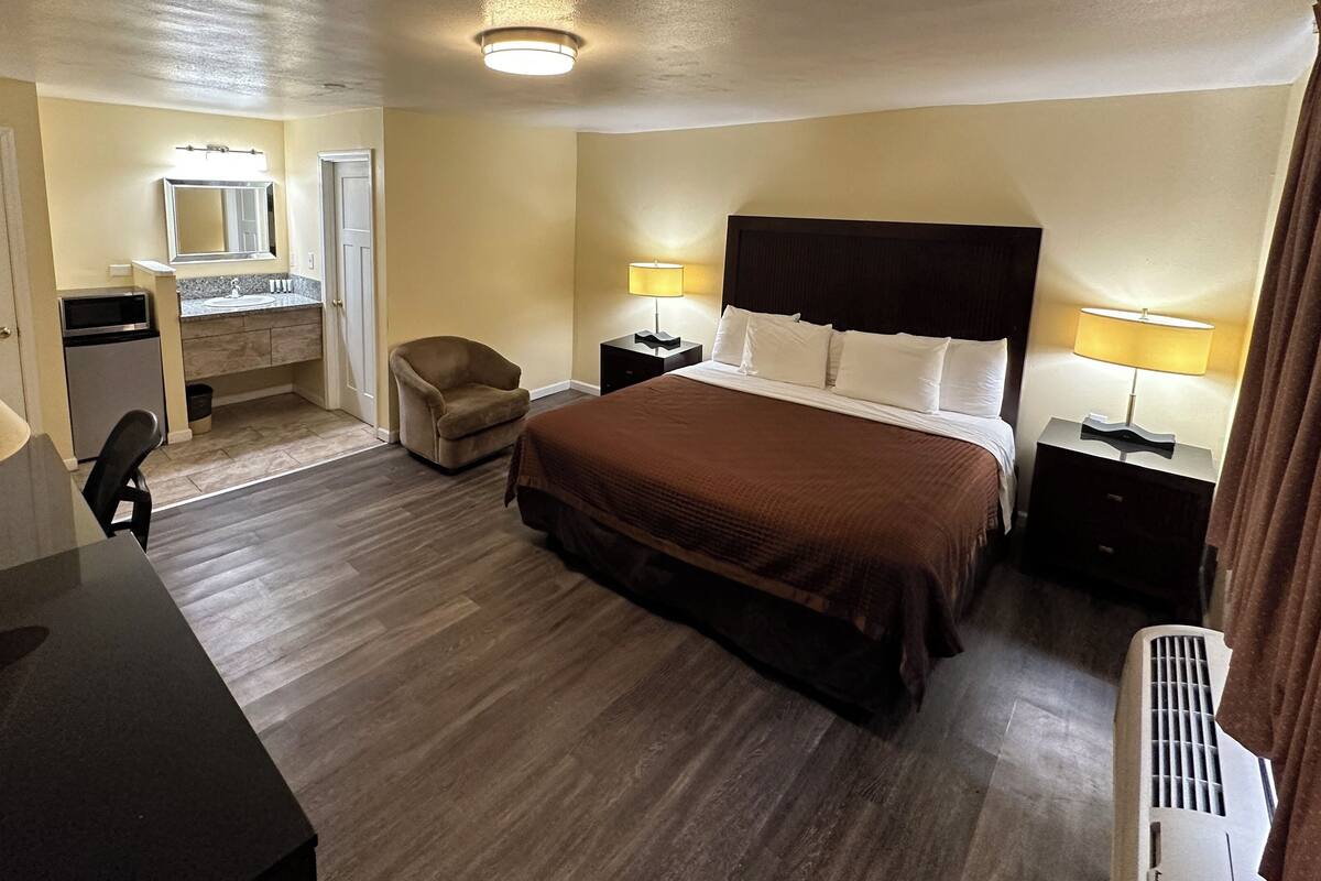 A hotel room with a bed, bedside tables, a desk, a chair, a mirror, a bathroom with a sink and mirror, and a heater.