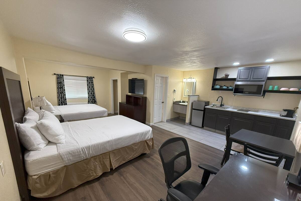 A compact hotel room with two beds, a kitchenette, a desk, and a chair.