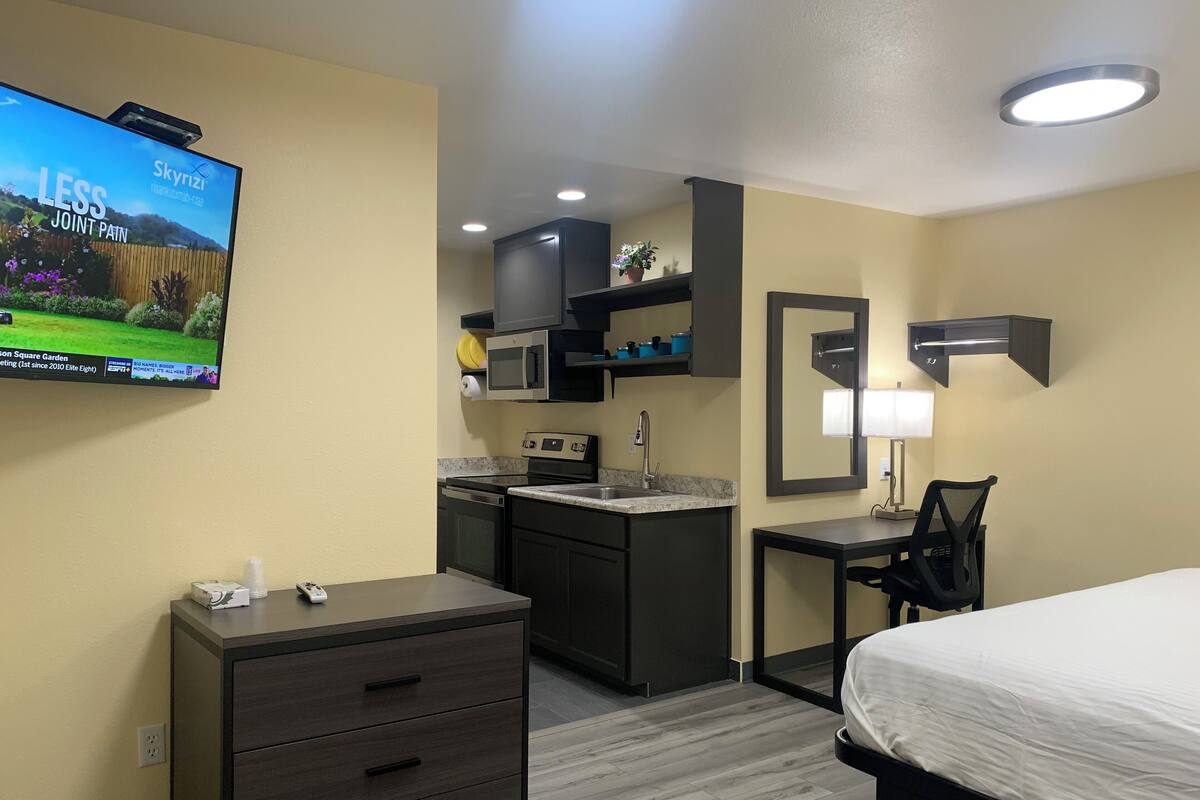 A hotel room with a bed, a desk with a chair, a kitchenette, a TV mounted on the wall, and a mirror.
