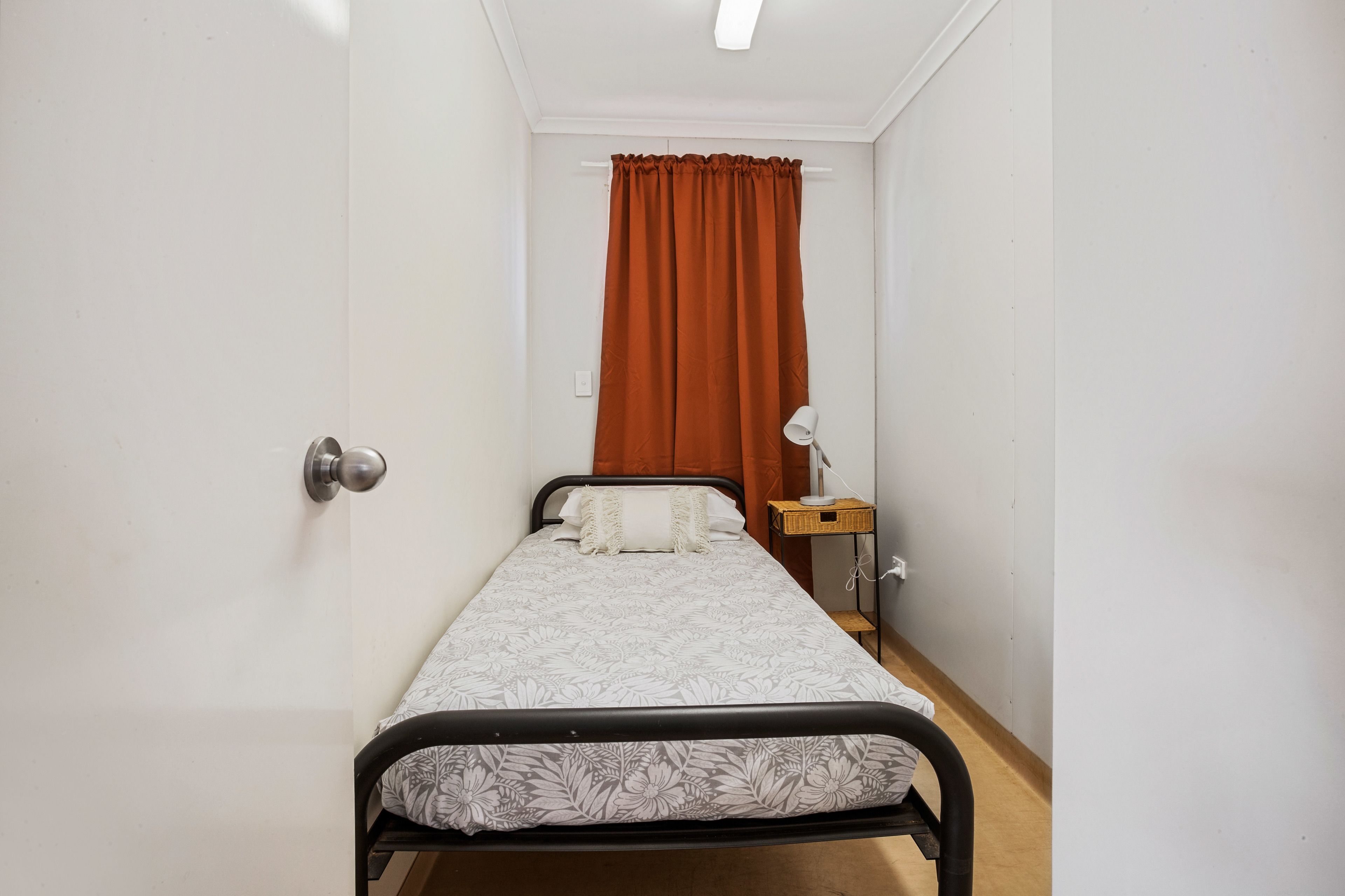 Et lille, pænt værelse med en seng, et natbord med en lampe og et vindue med orange gardiner.