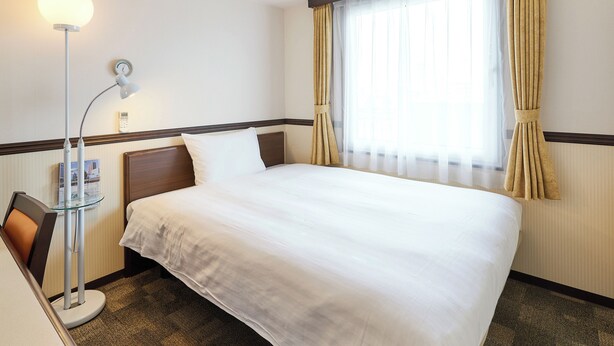 A hotel room with a neatly made bed, a bedside lamp, a clock on the wall, and a window with curtains. - Toyoko Inn Tsushima Izuhara (Tsushima)