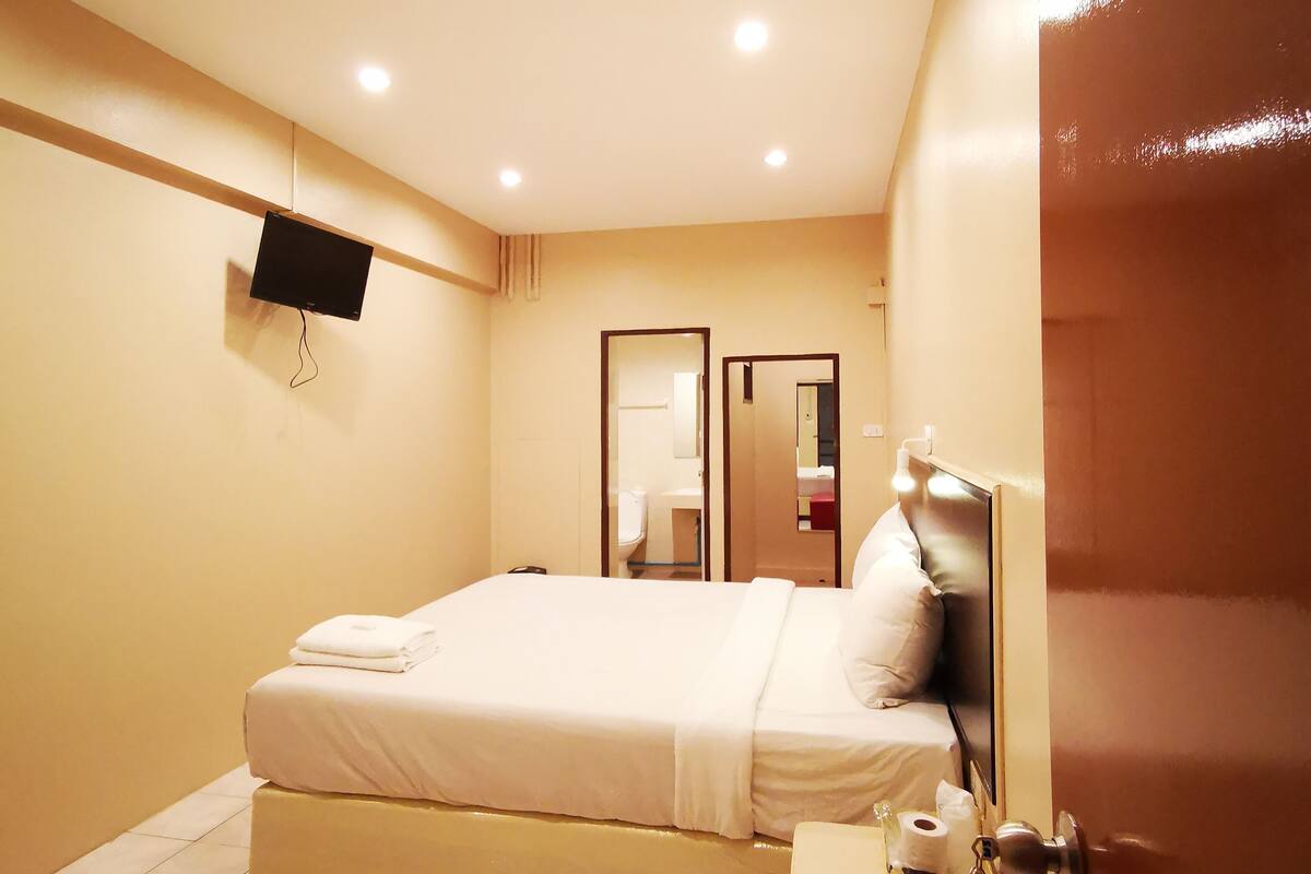 Une chambre d’hôtel avec un lit, une table de chevet avec des articles de toilette, un téléviseur fixé au mur et une porte munie d’une étiquette rouge.
