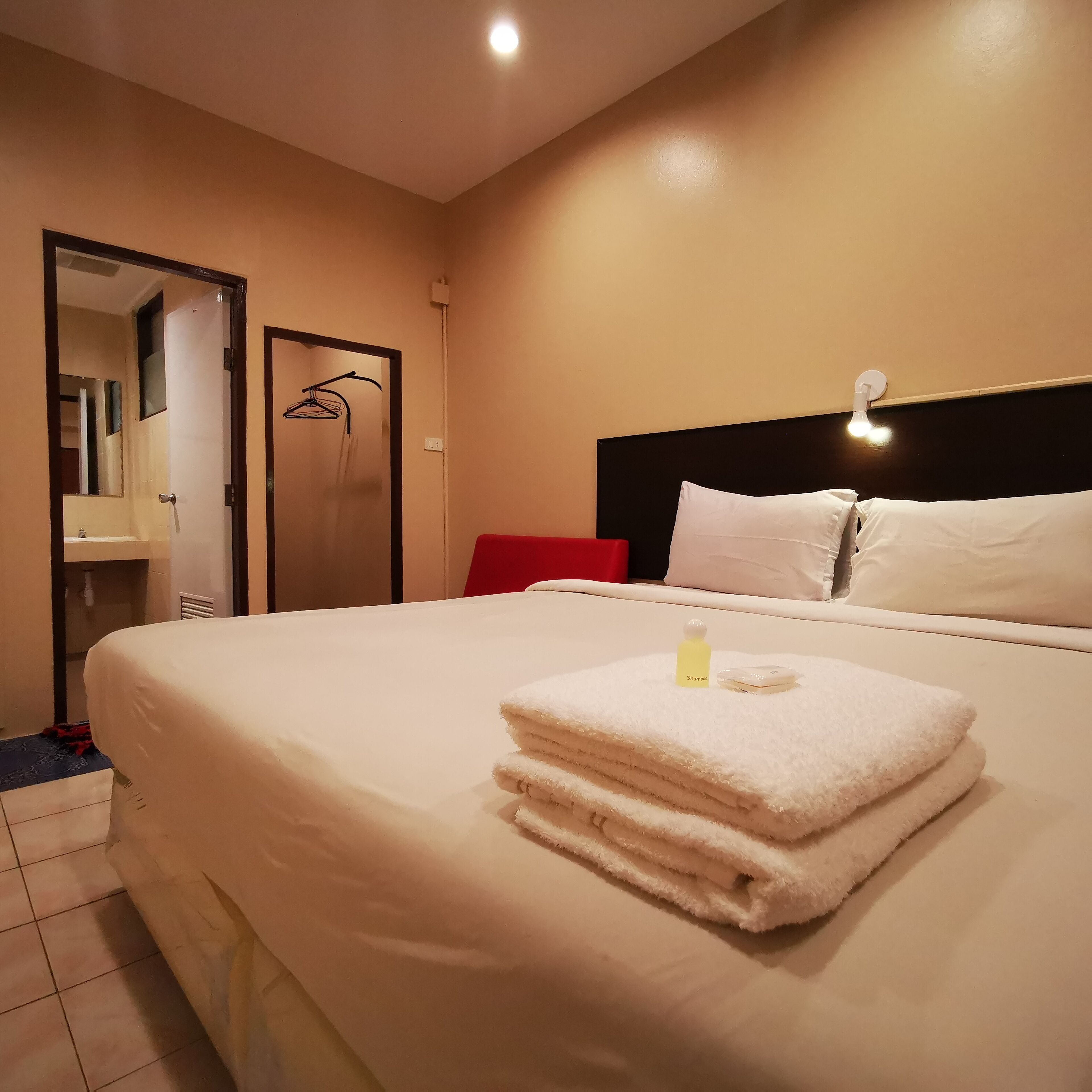 A hotel room with a neatly made bed, a red chair, a wall-mounted light, and a door leading to a bathroom.