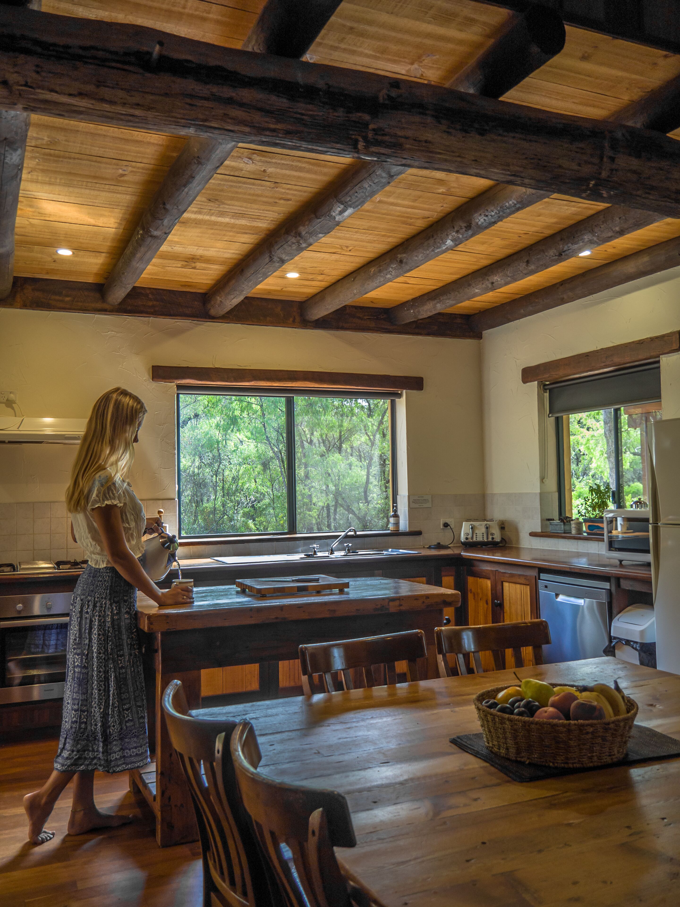 A rustic kitchen with wooden beams, a large window, and a dining area with chairs and a fruit basket.