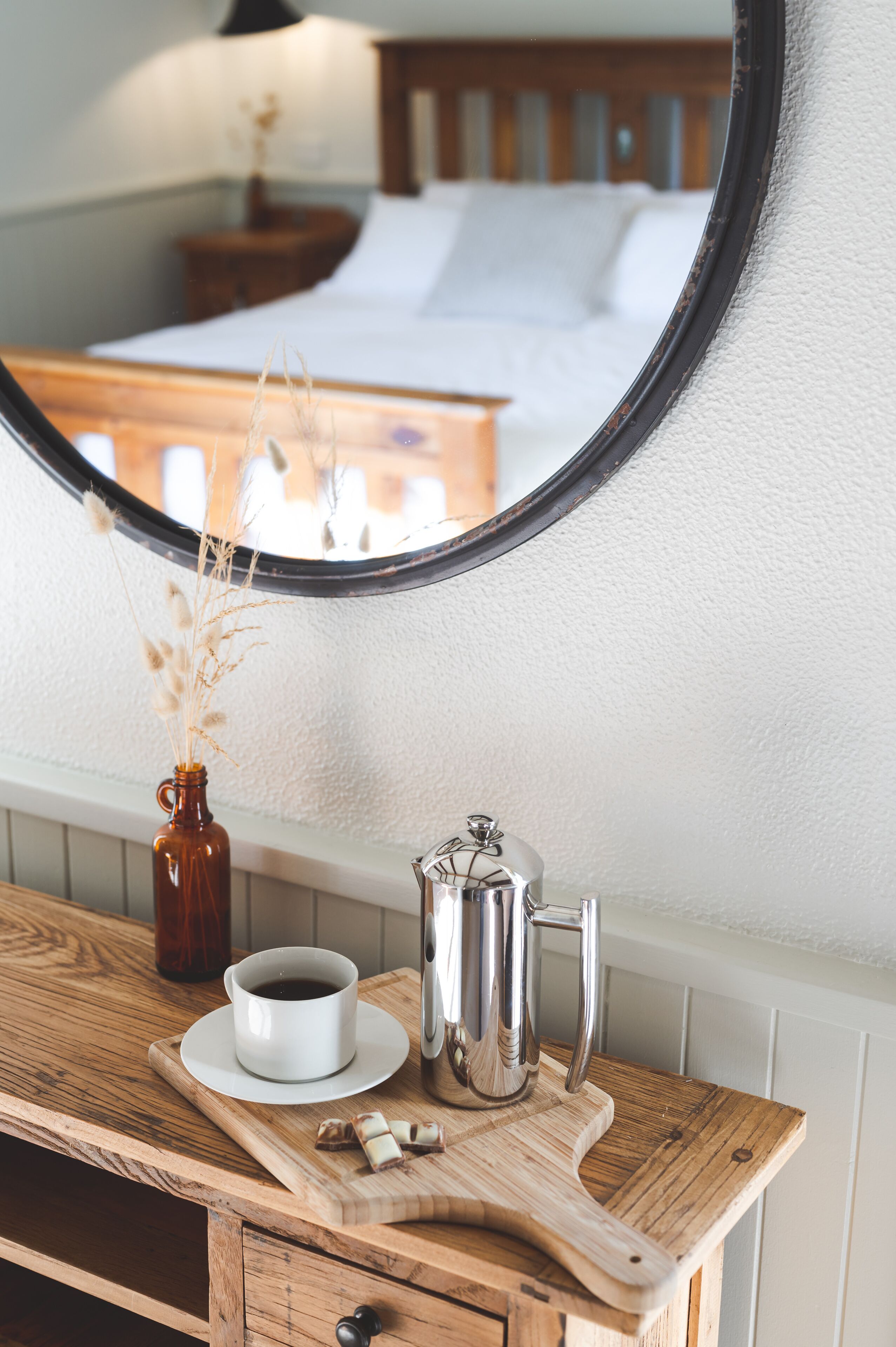 Une coiffeuse en bois avec un miroir, une tasse à café, un pichet en argent et un petit vase contenant des plantes séchées.