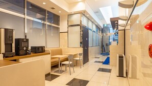 Un espacio de oficina moderno con divisiones de vidrio, una kitchenette, una zona de estar con sofá y sillas, y un bote de basura.