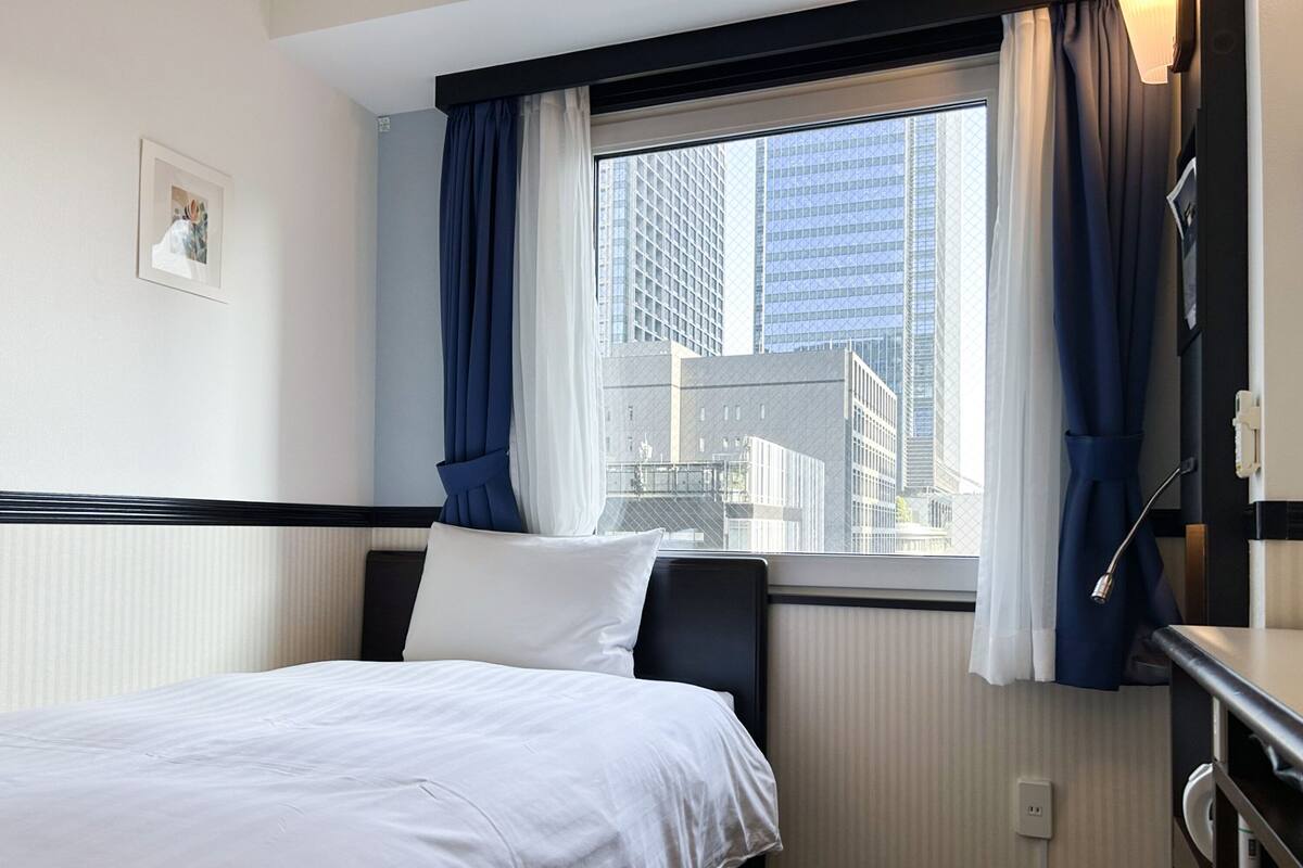 Une chambre d’hôtel avec un lit, une fenêtre avec des rideaux, un bureau et une vue sur de hauts immeubles.