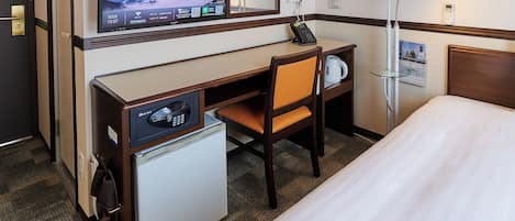 A hotel room with a bed, a desk with a computer monitor, a chair, a small refrigerator, a coffee maker, and a telephone.