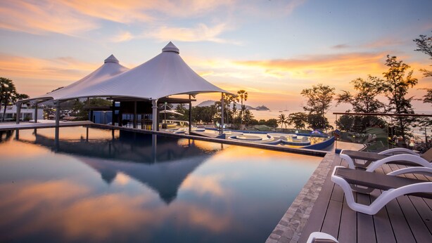 A serene pool area with two white canopies, a wooden deck, and a view of the sunset over the water.