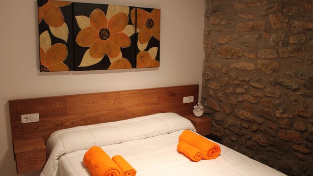 A neatly made bed with white linens and two rolled orange towels, a wooden headboard, a wall clock, and a large floral artwork above the bed. - Mountain apartment (SORT)