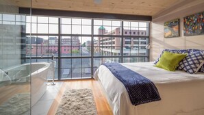 A modern bedroom with a large window, a bed with patterned bedding, a green pillow, and a white chair. - Old Port Penthouse Suite - Amazing Harbor Views (Portland)