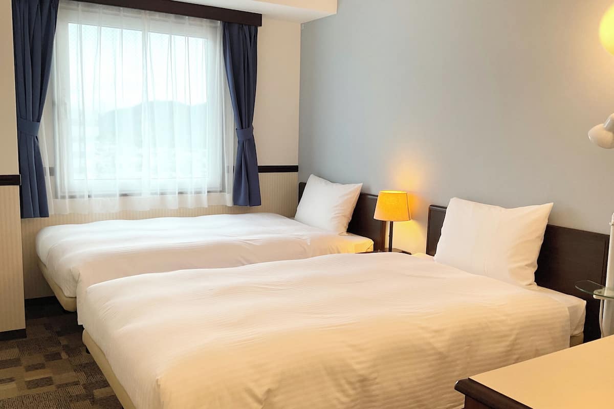 A hotel room with two beds, a window with curtains, a bedside lamp, and a small table.
