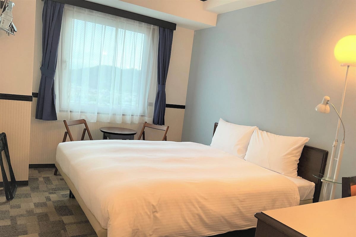 A hotel room with a large bed, a small table, a chair, a desk, a lamp, and a window with curtains.