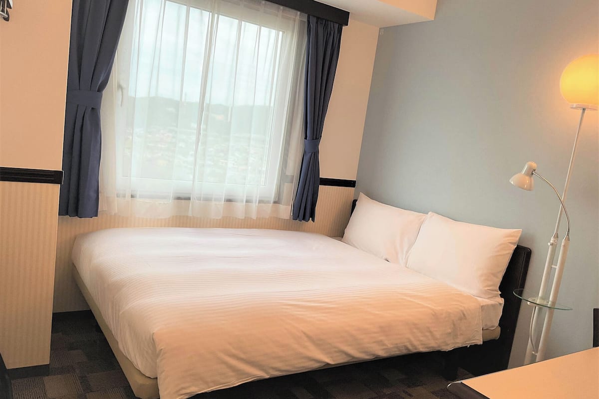 A hotel room with a bed, a window with curtains, a bedside lamp, and a wall-mounted shelf.
