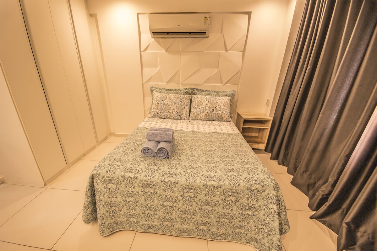 Una cama bien hecha con colcha y almohadas estampadas, un equipo de aire acondicionado sobre el cabecero y una cortina a la derecha.