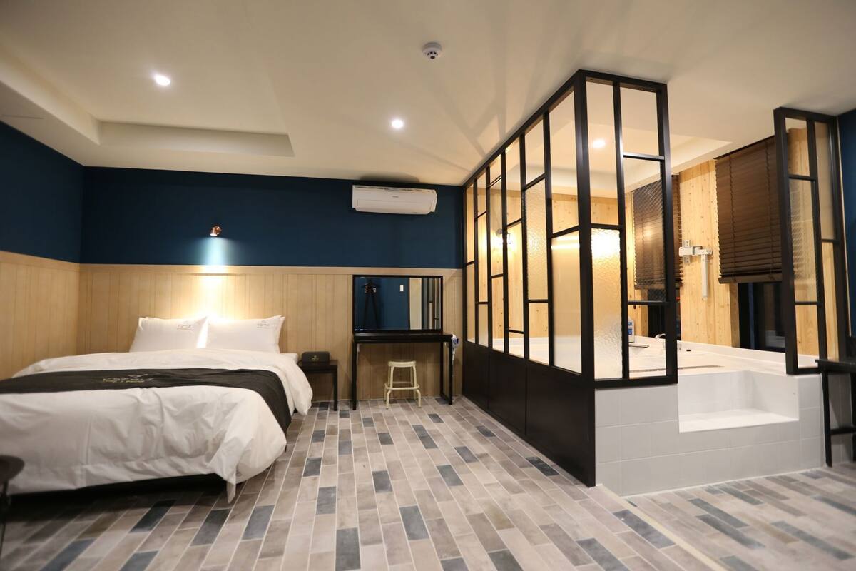 A hotel room with a bed, a desk, a chair, a bathroom with a bathtub, and a large window.
