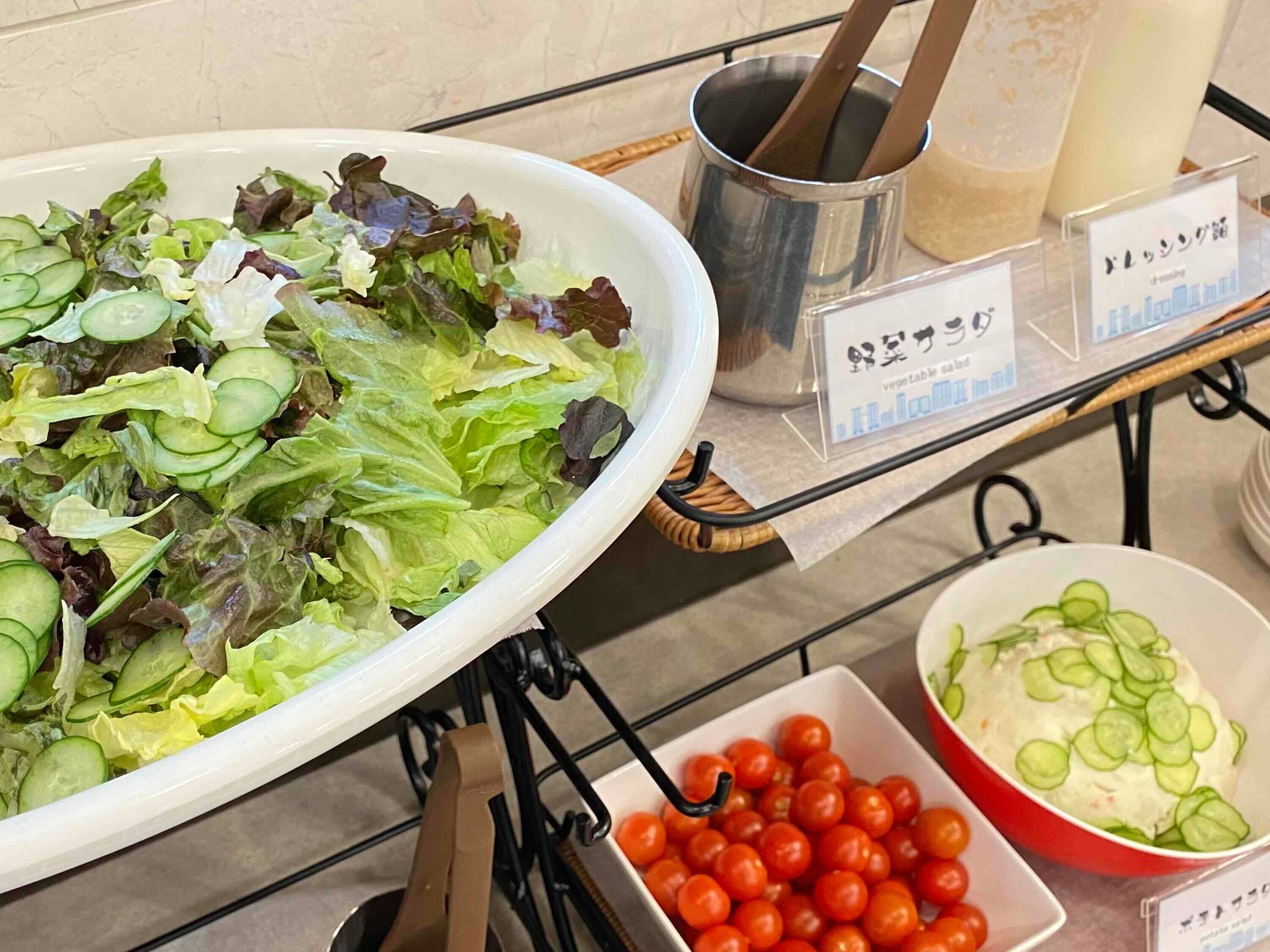Una mesa de buffet con un tazón grande de ensalada mixta y pepinos, un tazón de tomates cherry, y un recipiente con ensalada cremosa y rodajas de pepino.
