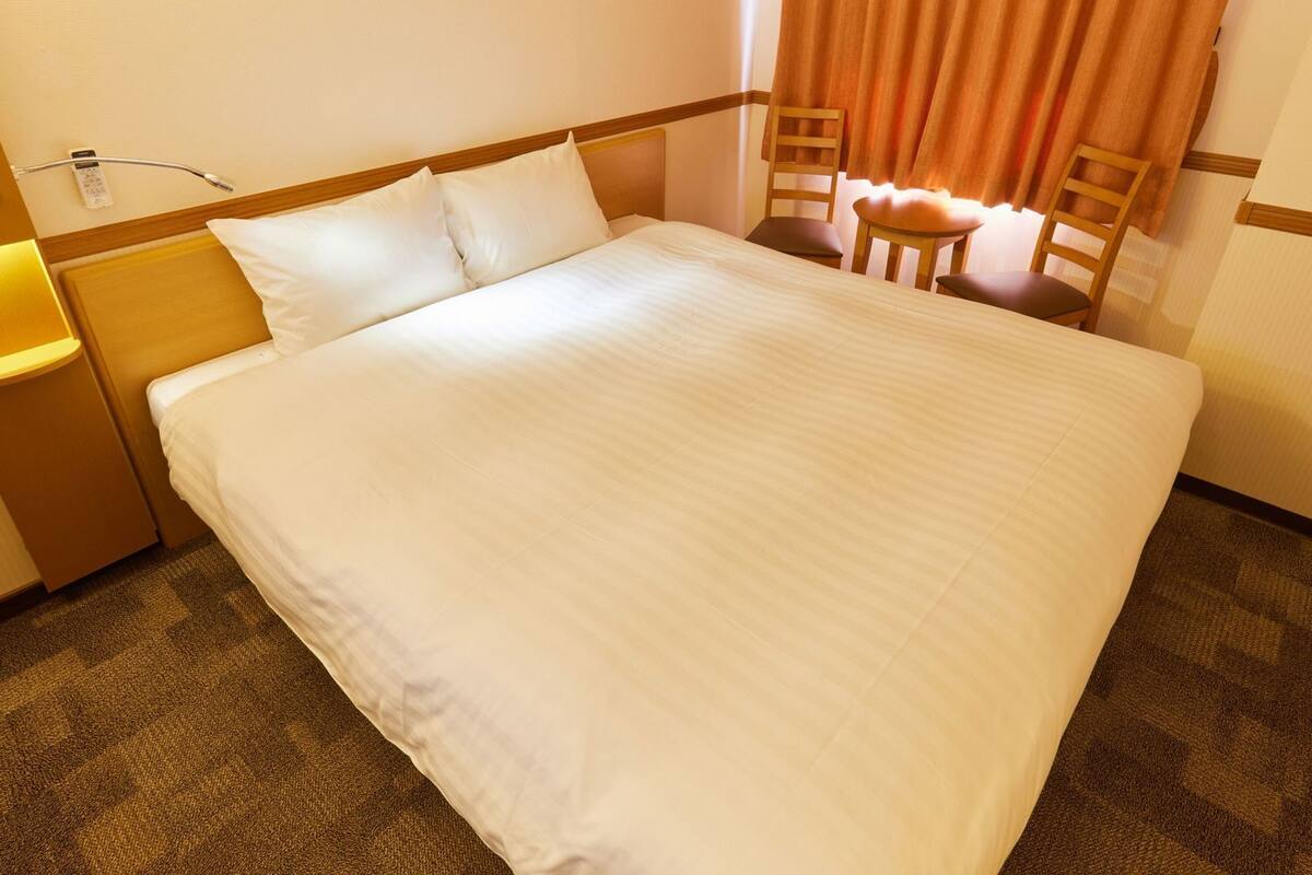 Une chambre d’hôtel avec un grand lit, une table de chevet, une lampe, deux chaises en bois et une fenêtre avec des rideaux.