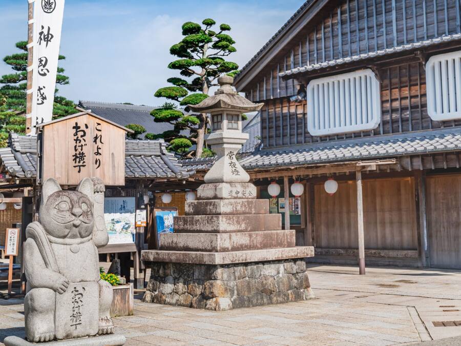 Um edifício japonês tradicional com um candeeiro de pedra, uma estátua de gato de pedra e uma placa com caracteres japoneses.