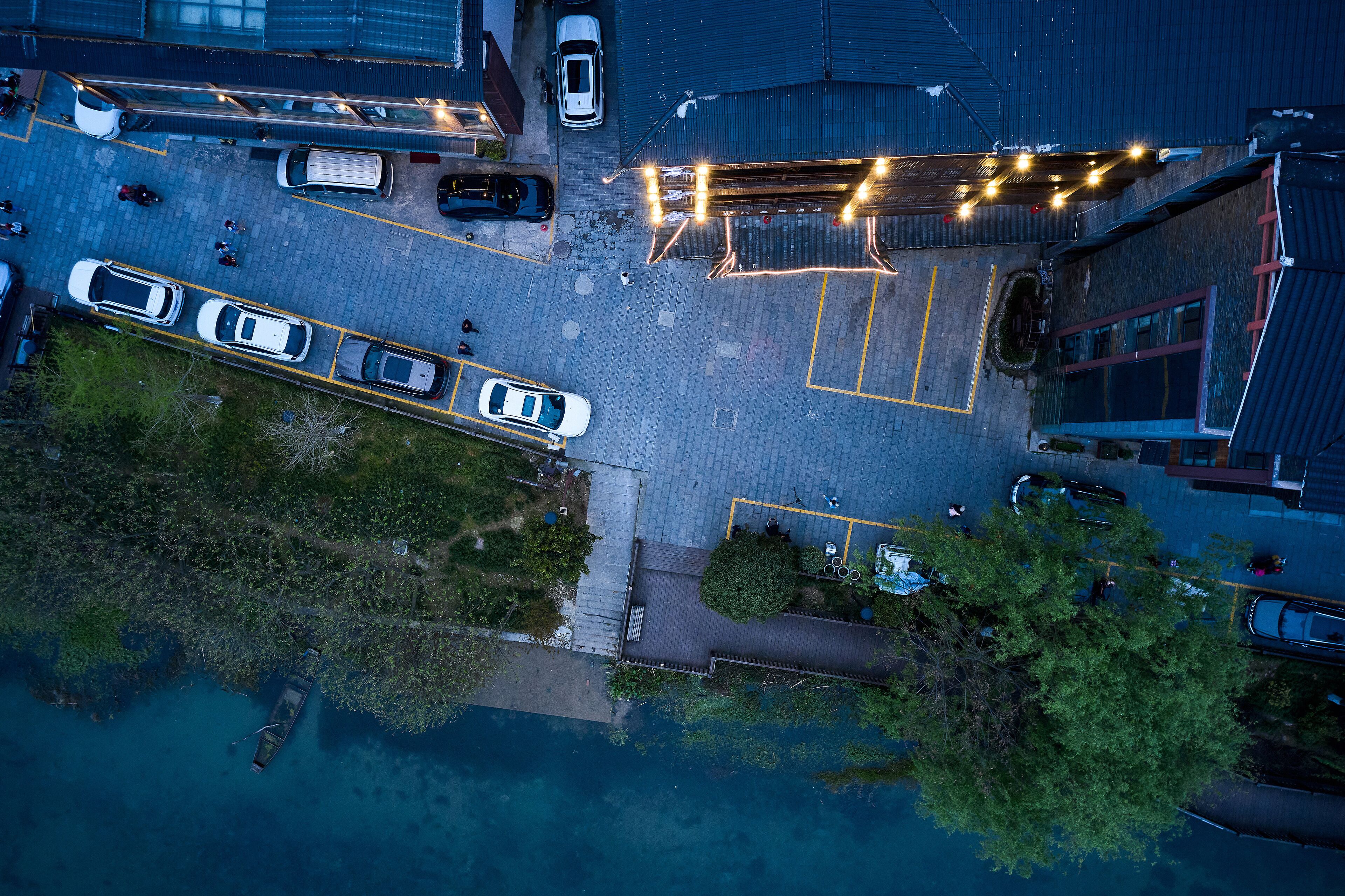 An aerial view of a parking lot with several cars, a boat docked near a body of water, and a building with a sloped roof.