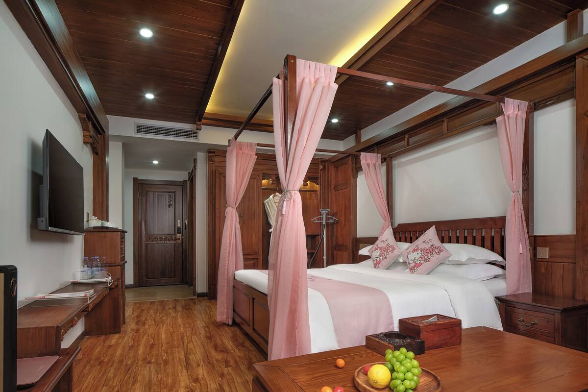 Un dormitorio con techo de madera, una cama con cortinas rosas, una mesita de noche, un televisor y un frutero sobre una mesa de madera.