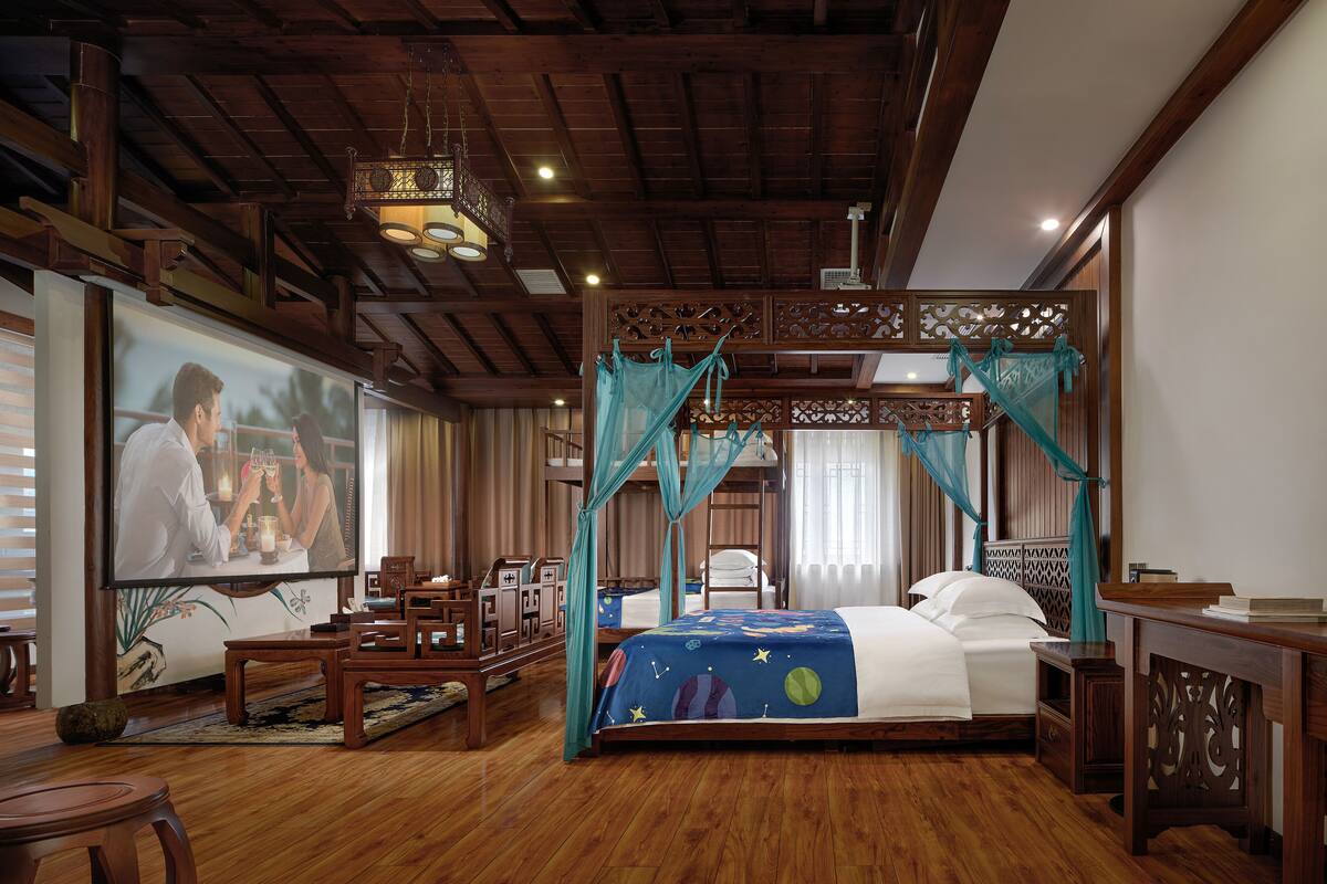 Una habitación tradicional con techo de madera, una cama con cubrecama azul, un escritorio de madera y una televisión mostrando una pareja.