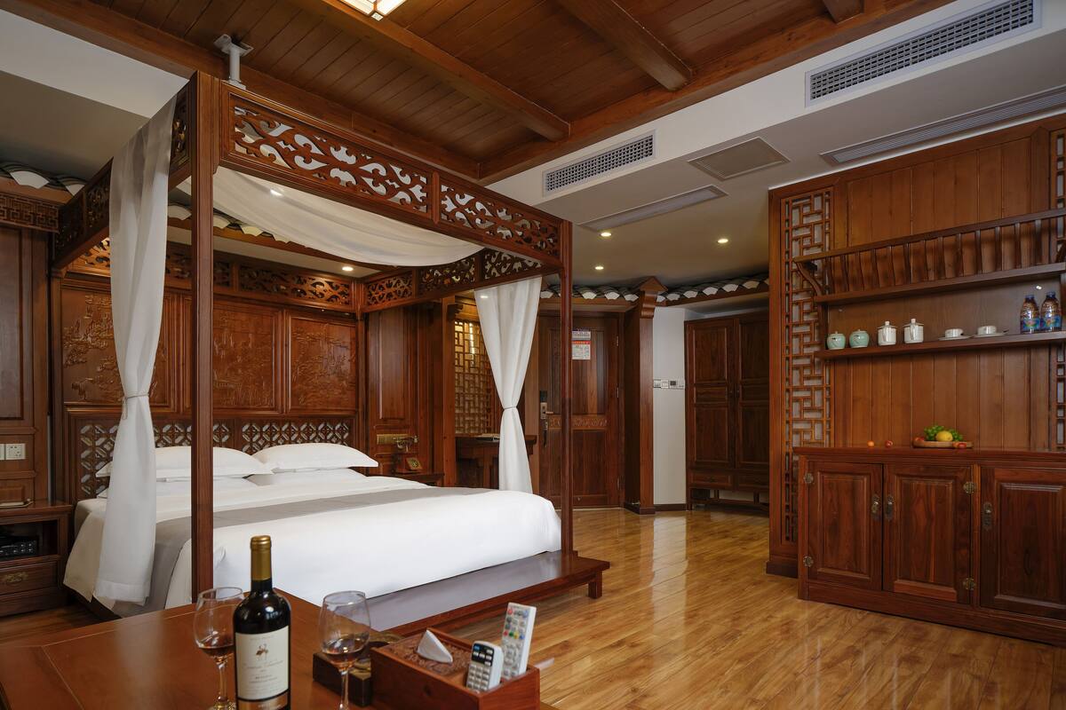 Un dormitorio tradicional de madera con cama con dosel, un armario de madera y una mesa de madera con una botella de vino y una copa.