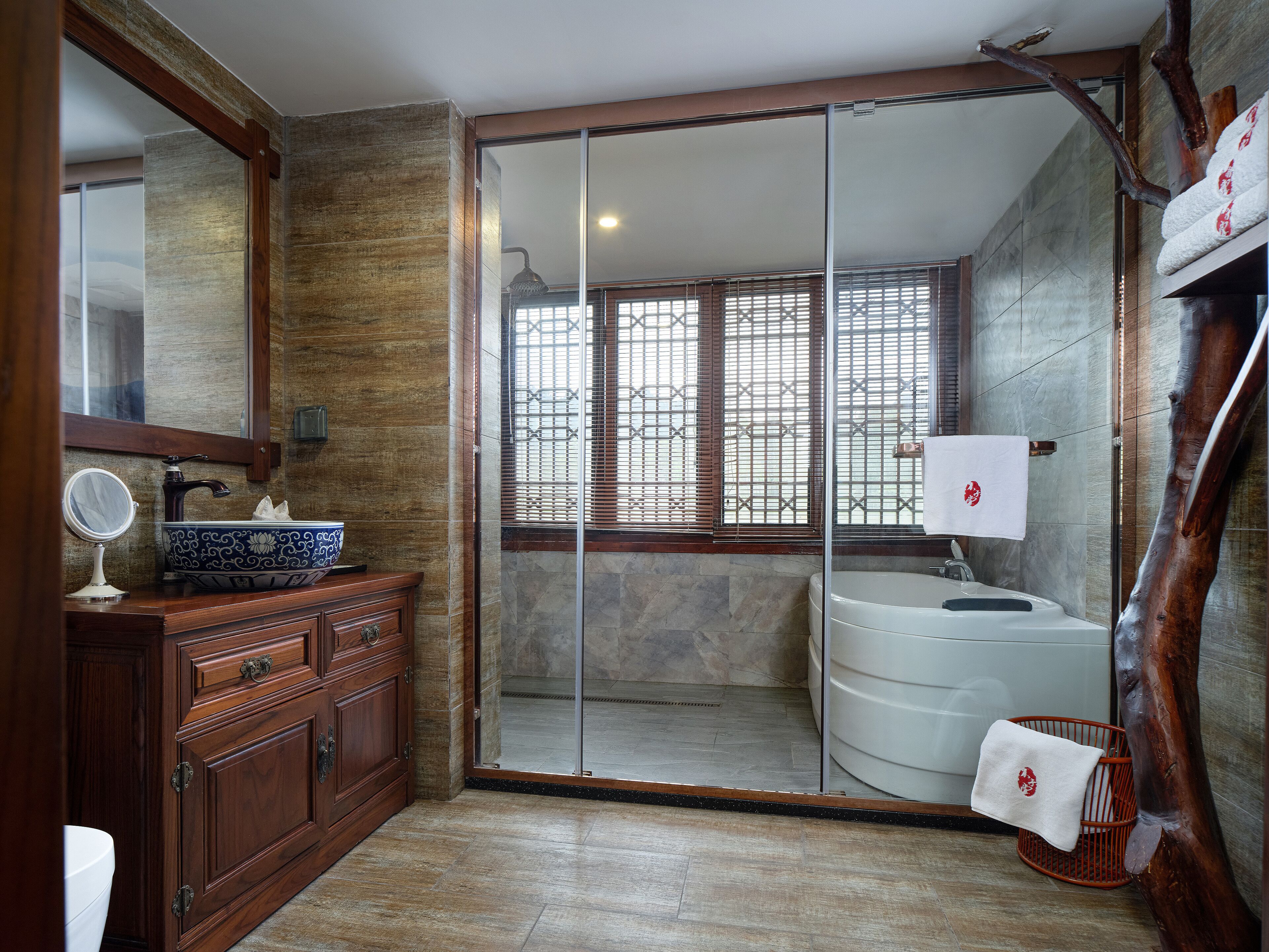 A bathroom with a wooden vanity, a round mirror, a white sink with a decorative bowl, a large mirror above the vanity, a shower with a glass door, and a towel rack with a towel.