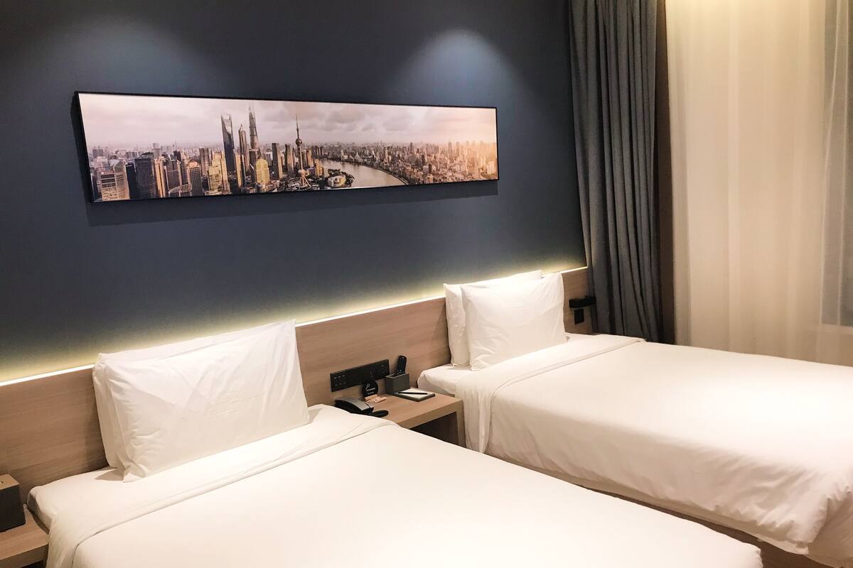 A hotel room with two beds, a nightstand, a remote control, and a cityscape painting on the wall.
