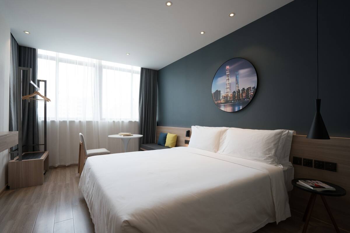 A modern hotel room with a large bed, a round cityscape painting on the wall, a bedside table, and a chair.