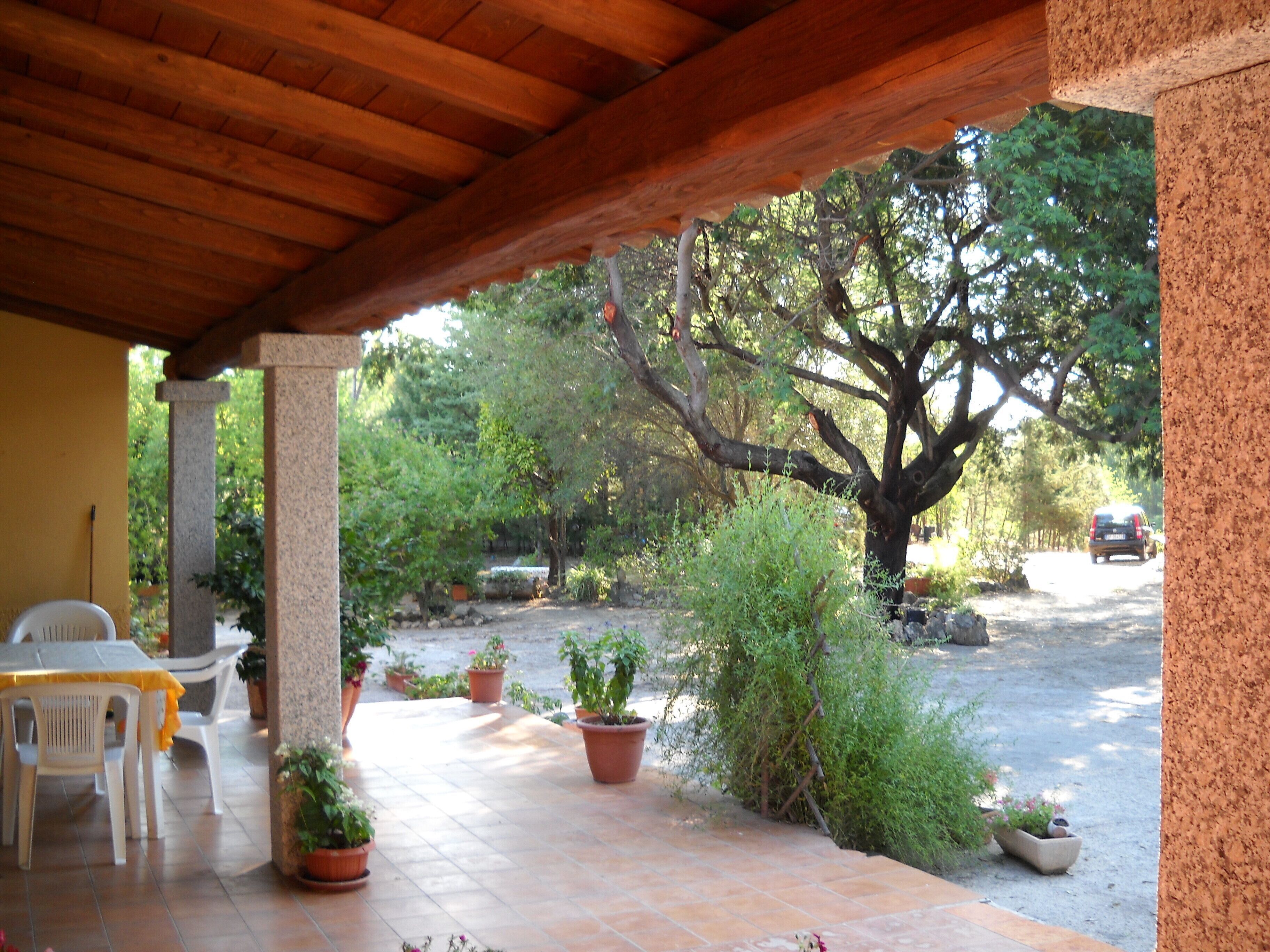 A covered patio area with a table and chairs, surrounded by potted plants and a view of a tree-lined street.