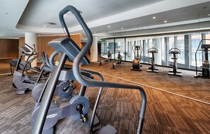 Un gimnasio con diversas máquinas de ejercicio, piso de madera y ventanas amplias con vistas.