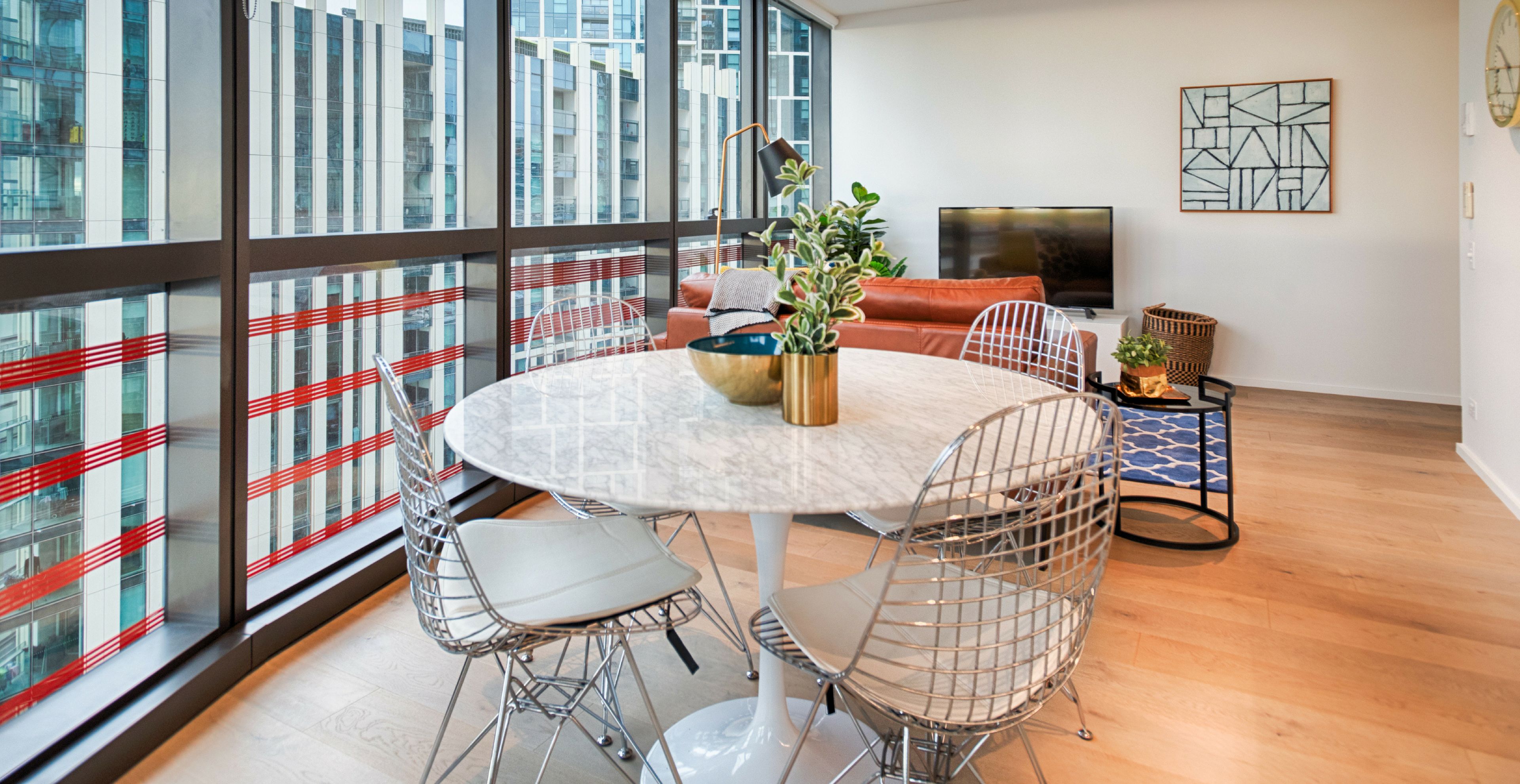 A modern dining area with a round table, metal chairs, and a view of a cityscape through large windows.