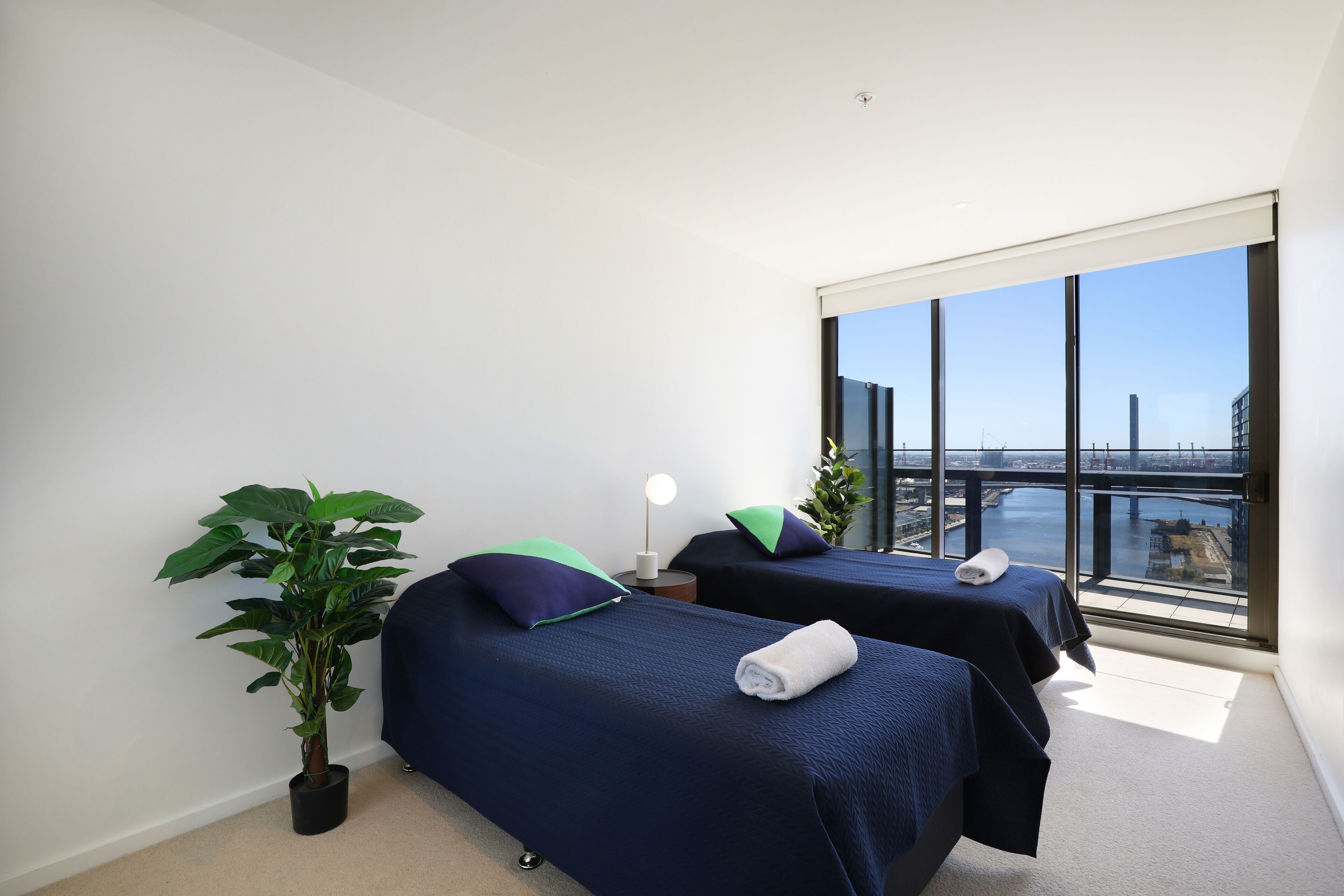 A hotel room with two beds, a potted plant, a nightstand with a lamp, and a large window offering a city view.