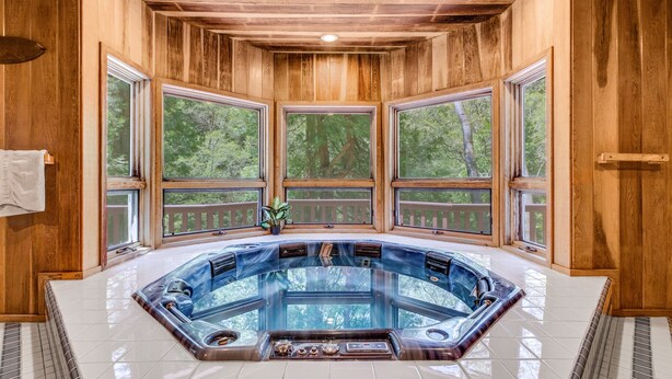 A wooden cabin with a hot tub in the center, large windows, and a view of trees.