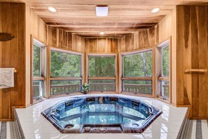 Une cabane en bois dotée d’un jacuzzi au centre, de grandes fenêtres et offrant une vue sur les arbres.