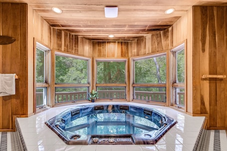 A wooden cabin with a hot tub in the center, large windows, and a view of trees.
