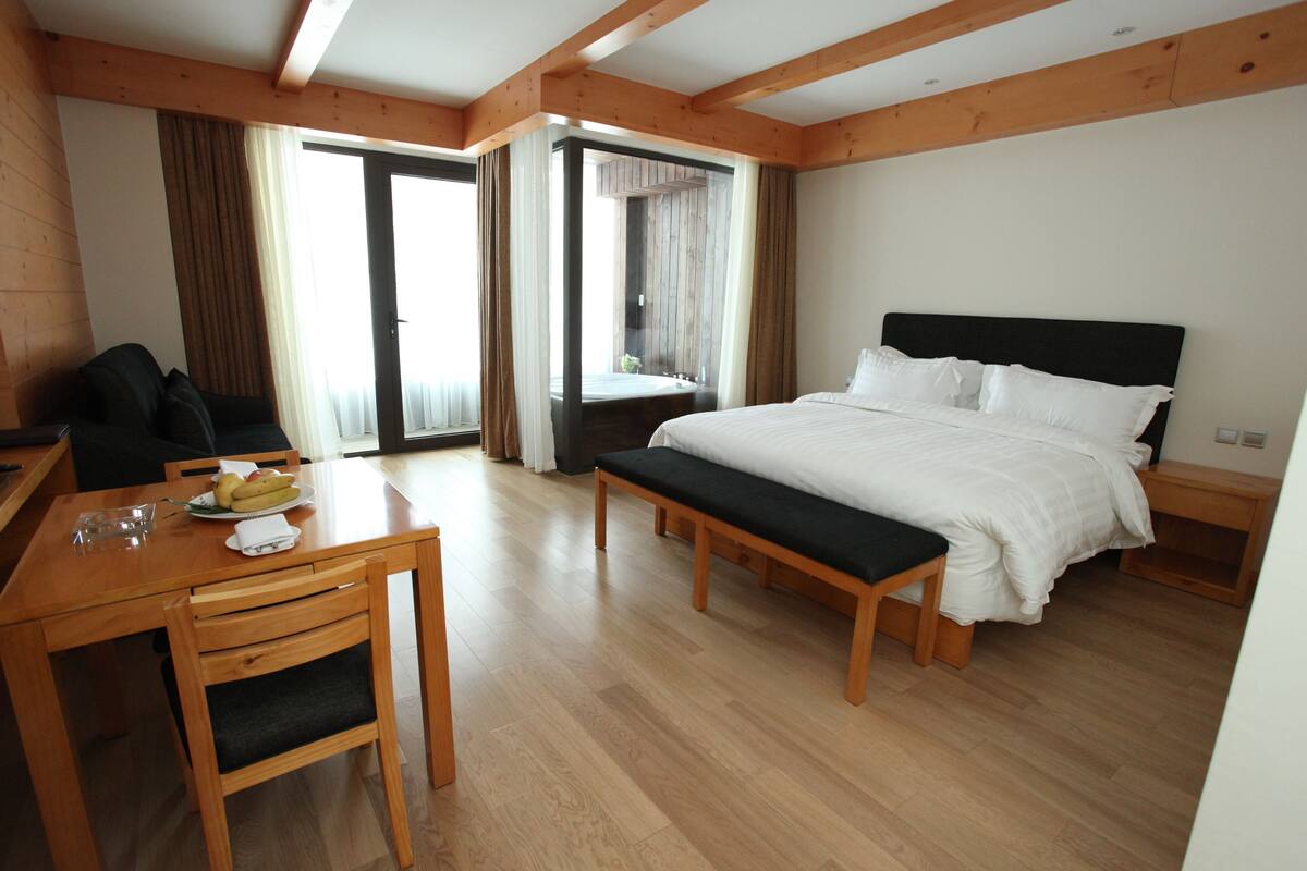 A hotel room with a wooden floor, a bed with white bedding, a bench, a desk with a chair, and a dining table with a plate of fruit.