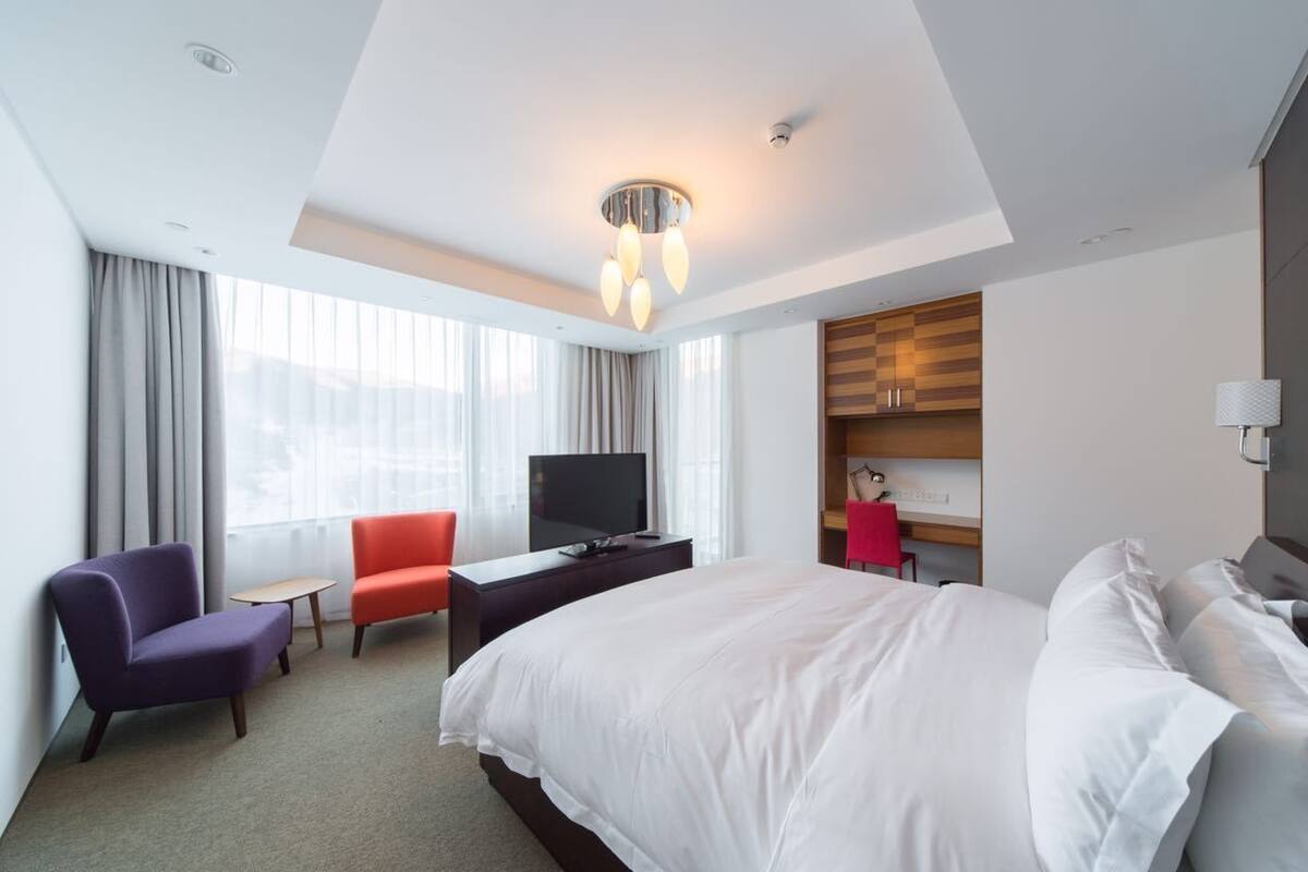 A hotel room with a large bed, a desk with a TV, two chairs, a small table, and a wardrobe.